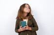 © Jihan - Portrait of Beautiful little girl standing against white background, with dreamy look, thinking while holding smartphone. Tries to write up a message.