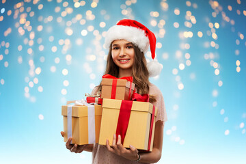 Wall Mural - christmas, holidays and people concept - happy smiling teenage girl in santa helper hat holding gift box over lights on blue background