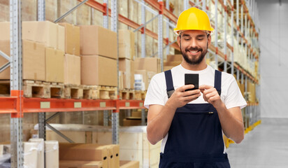 Canvas Print - logistics, profession and job concept - happy smiling male worker or loader in yellow helmet and overall with smartphone over warehouse background