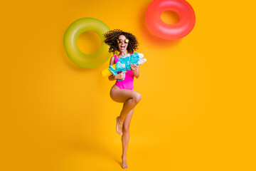 Wall Mural - Photo portrait of cheerful young brunette girl standing on one leg holding water gun laughing wearing fuchsia swim wear isolated on vivid yellow colored background with green red inflatable rings