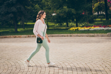 Poster - Full size profile photo of cute brunette lady walk park active life wear pastel green jeans white top striped shirt shoulder bag outdoors