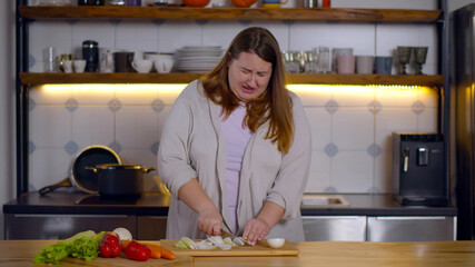 Portrait of overweight woman on diet crying chopping onion in kitchen