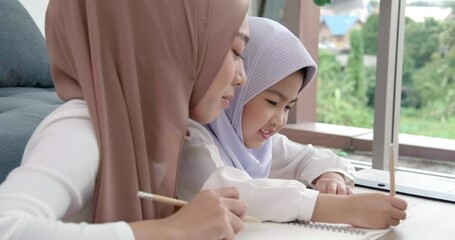 Beautiful young traditional Islamic muslim Asian mother in house life style working teach her daughter education homework from laptop PC on the wooden table and writing to book