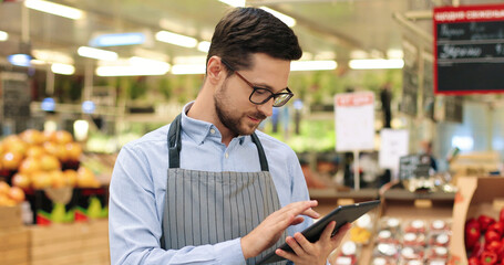 Close up portrait of happy Caucasian male worker in glasses standing in supermarket and typing on tablet. Young joyful man food store assistant at work tapping on device indoors. Retail concept