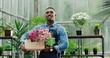 © VAKSMANV - Portrait of happy handsome ypung African American businessman standing in apron in own flower shop and smiling to camera while holding floral box. Joyful male in green house at work. Floristry concept