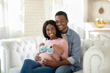 Black Pregnant Woman With Tiny Baby Boties And Her Happy Husband Hugging On Sofa At Home