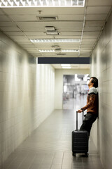 Wall Mural - Asian man tourist with face mask feeling exhausted leaning on wall while carrying suitcase luggage in airport terminal. Coronavirus (COVID-19) pandemic prevention when travel. Health insurance concept