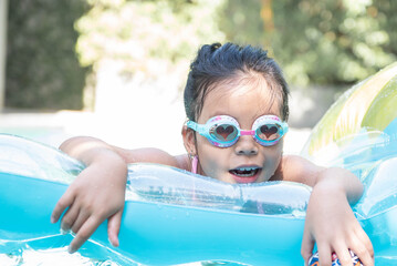 Wall Mural - happy little girl playing with inflatable ring in outdoor swimming pool on hot summer day. kids learn to swim. child water toys. children play in tropical resort. family beach vacation.