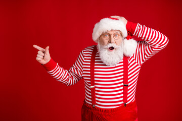 Wall Mural - Photo of retired old man grey beard direct finger empty space hand head shocking baubles sale wear santa x-mas costume suspenders spectacles striped shirt cap isolated red color background