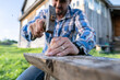 © koldunova - Focused male carpenter makes a bench hammering nails into a board in a village courtyard.