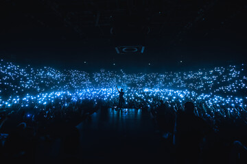 Vocalist in front of crowd on scene in 
stadium. Bright stage lighting, crowded dance floor. Phone lights at concert. Band blue silhouette crowd. People with cell phone lights.