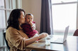 Mom holding baby and working from home on laptop at dining room table 