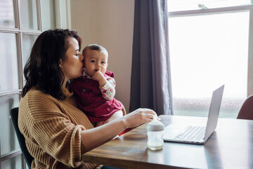 Mom holding baby and working from home on laptop at dining room table 