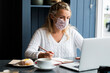 © Mint Images - Woman wearing face mask sitting alone at a cafe table with a laptop computer