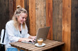 © Mint Images - Young blond woman sitting alone at a cafe table with a laptop computer, working remotely.