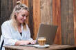 © Mint Images - Young blond woman sitting alone at a cafe table with a laptop computer, working remotely.