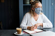 © Mint Images - Woman wearing face mask sitting alone at a cafe table with a notebook and pencil