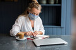 © Mint Images - Woman wearing face mask sitting alone at a cafe table with a note book and calculator, working remotely.