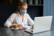 © Mint Images - Woman wearing face mask sitting alone at a cafe table using a laptop working remotely.