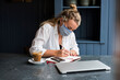 © Mint Images - Woman wearing face mask sitting alone at a cafe table writing in a notebook working remotely.