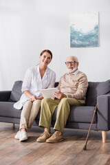 Wall Mural - cheerful aged man holding digital tablet near smiling social worker while sitting o sofa at home