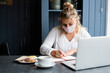 © Mint Images - Woman wearing face mask sitting alone with a laptop, writing in note book, working remotely.