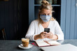 © Mint Images - Woman wearing face mask sitting alone at a cafe table with a laptop computer, using mobile phone