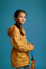 Wall Mural - cute little child girl with multicolored umbrella in yellow rain coat on blue background. autumn mood