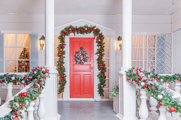 Christmas porch decoration idea. House entrance with red door decorated for holidays. Red and green wreath garland of fir tree branches and lights on railing. Christmas eve at home