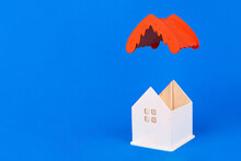 Wooden Model Of A House Without A Roof On A Blue Background. The Red Roof Of The House Flying From Above. Cover The Standing House With A Roof. Isolate. Empty Space For Design. Copy Space