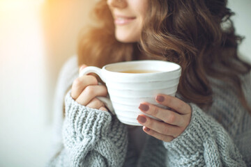 Close-up of female hands with a mug of drink. Beautiful girl in a gray sweater is holding a cup of tea or coffee in the morning sunlight. Mug for your design. High quality photo.