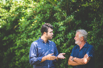 an adult hipster son and senior father talking at home, morning nature scene in the home garden outd