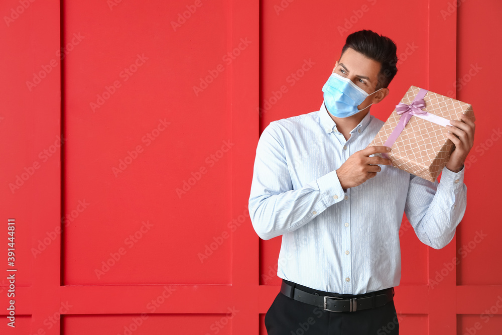 Young man in medical mask and with gift on color background