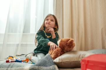 Wall Mural - A small girl playing doctor with a toy stethoscope having a teddybear as a patient deciding what to do while sitting on a sofa in a spacy room