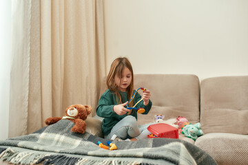 A small girl holding a toy stethoscope while sitting on a sofa with a teddybear and plaid near to her