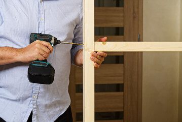 Wall Mural - A man fastens two wooden beams with a self-tapping screw. The joiner wraps the screw with a screwdriver. Construction of a timber frame for plasterboard