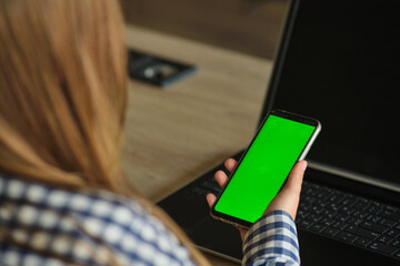 Wall Mural - Girl with glasses holding a phone with a green screen sitting at a laptop.