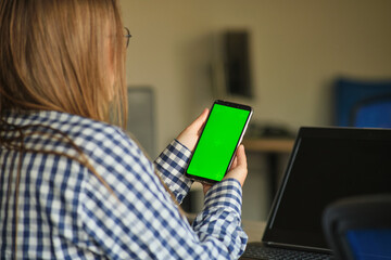 Wall Mural - girl with glasses holding a phone with a green screen sitting at a laptop.