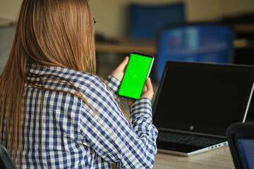 Wall Mural - girl with glasses holding a phone with a green screen sitting at a laptop.