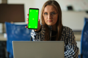 Wall Mural - Girl with glasses holding a phone with a green screen sitting at a laptop.