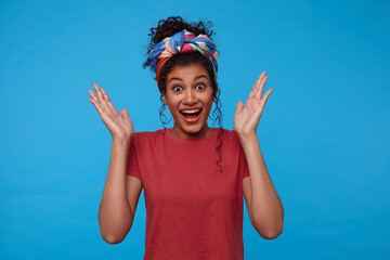 Wall Mural - Surprised young lovely brown haired female with gathered hair looking excitedly at camera and keeping hands raised while standing against blue background