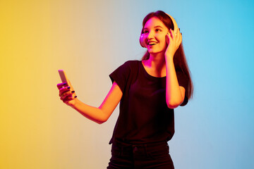 Wall Mural - Listening to music with headphones. Young caucasian woman's portrait on gradient blue-yellow studio background in neon. Concept of youth, human emotions, facial expression, sales, ad. Beautiful model.