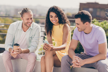 Photo of three positive fellow friends sitting look phone screen funny video drink coffee gathering outdoors