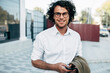© iuricazac - Confident businessman in eyewear smiling broadly posing outdoors. Male entrepreneur messaging on smartphone. Smart guy in casual wears spectacles with curly hair walking outside after work