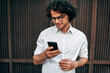 © iuricazac - Closeup of happy businessman in white shirt with transparent eyeglasses messaging on mobile phone. Handsome curly male has online conference with a colleague via smartphone outside.