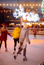 Young Woman Ice Skating On A Rink In A Festive Christmas Fair In The Evening. Smiling Woman In Winter Style Clothes Skates.  Winter Holidays Concept. Lights Around.