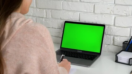 Green screen laptop computer concept: online distance education by web cam video call conference. Over the shoulder woman student at home study writes information in notebook listens teacher tutor