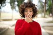 © Roquillo - Emotional Young arab woman wearing casual red sweater in the street gasps from astonishment, covers opened mouth with palm, looks shocked at camera. Reaction concept