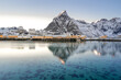 Beautiful winter fishing town on Nordic shore with tall snow mountain backdrop and reflection on the turquoise water. overcast  sky and clouds and evening serene light in cold lonely winter season. 