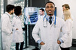 © sofiko14 - Young handsome smiling african american medical doctor with his multiracial colleagues, standing on the background, having video chat with male doctor and duscussing patient's CT